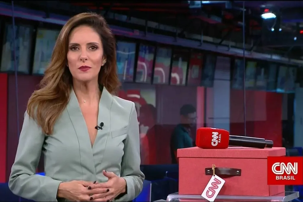 Jornalista Monalisa Perrone de blazer verde água, com as mãos cruzadas à frente do corpo, em pé no estúdio da CNN Brasil. Ao lado dela, sobre uma bancada, há uma caixa vermelha com um microfone da emissora em cima.