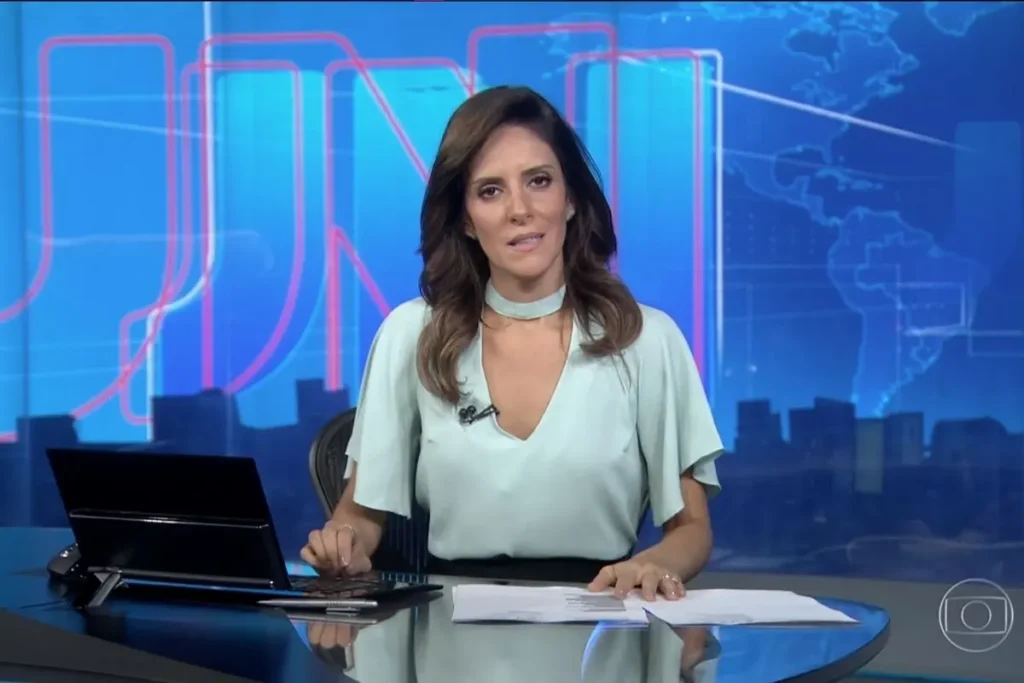 Jornalista Monalisa Perrone sentada à bancada do Jornal Nacional, vestindo blusa verde clara. Ao fundo, o cenário azul característico do telejornal com o logotipo JN em linhas vermelhas e um mapa-múndi estilizado.