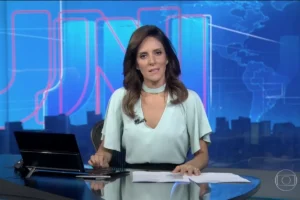 Jornalista Monalisa Perrone sentada à bancada do Jornal Nacional, vestindo blusa verde clara. Ao fundo, o cenário azul característico do telejornal com o logotipo JN em linhas vermelhas e um mapa-múndi estilizado.