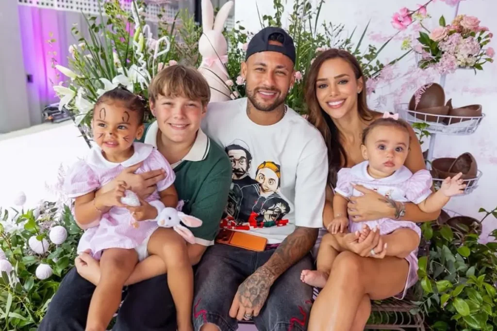 Neymar Jr., usando boné preto virado para trás, Bruna Biancardi, o filho mais velho Davi Lucca, de camisa polo verde, e as duas filhas pequenas, Mavie e Helena, ambas com vestidos rosa e branco, posam juntos. Mavie está com o rosto pintado de coelho. O grupo está cercado por decoração de Páscoa com ovos de chocolate e arranjos florais de tons rosa.