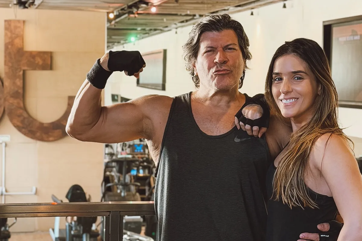 O cantor Paulo Ricardo posa ao lado de sua namorada, Isabella Pinheiro, em uma unidade da academia Bodytech. Paulo Ricardo, vestindo regata cinza, faz uma pose de "muque" exibindo o bíceps e usa luvas de treino pretas. Isabella sorri, abraçada a ele, vestindo roupa esportiva preta. Ao fundo, vê-se o logotipo "bt" em destaque na parede e aparelhos de musculação.