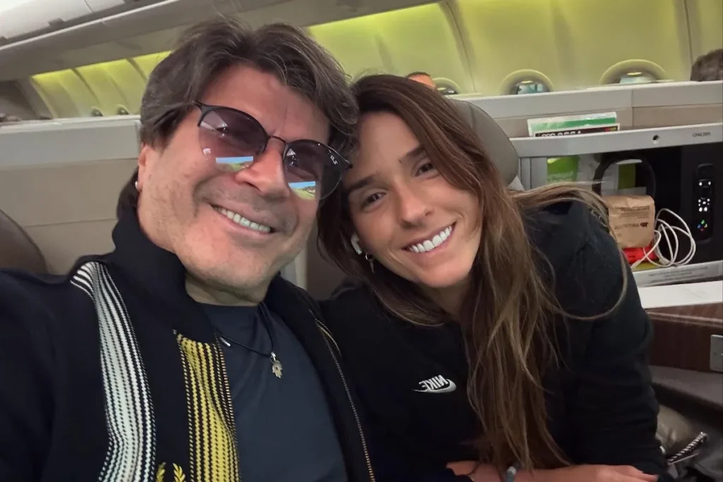 O cantor Paulo Ricardo e sua namorada, Isabella Pinheiro, sorriem em uma selfie tirada a bordo de um avião. Paulo Ricardo, à esquerda, usa óculos escuros de sol e uma jaqueta preta com detalhes em amarelo e branco sobre uma camiseta preta. Isabella, à direita, veste um moletom preto da Nike com o logo branco visível e tem o cabelo comprido solto. O ambiente mostra o interior da cabine da classe executiva da LATAM, com assentos largos e as laterais verdes da aeronave visíveis.