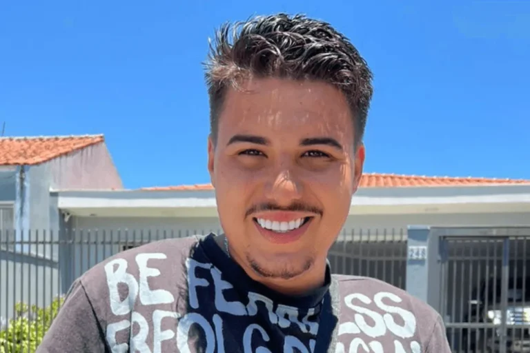 Pedro, do BBB 26. Homem jovem sorridente de pele clara, com cabelos escuros curtos e levemente espetados, bigode e cavanhaque finos. Ele veste uma camiseta cinza com letras brancas e está em frente a uma casa branca com portão cinza, sob um céu azul limpo.