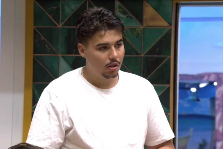 Pedro Espíndola, do BBB 26. Homem jovem com bigode, cavanhaque e cabelo castanho cacheado, vestindo uma camiseta branca de algodão, falando com a boca ligeiramente aberta, em um cenário interno com fundo de parede de azulejos verdes e dourados e uma janela com vista para a paisagem.
