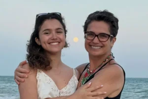 Sandra Annenberg e sua filha, Elisa Paglia, sorriem abraçadas em uma praia durante o entardecer. Sandra usa óculos de grau e um colar colorido, enquanto Elisa veste uma blusa branca de renda. Ao fundo, o mar e uma lua cheia no céu claro.