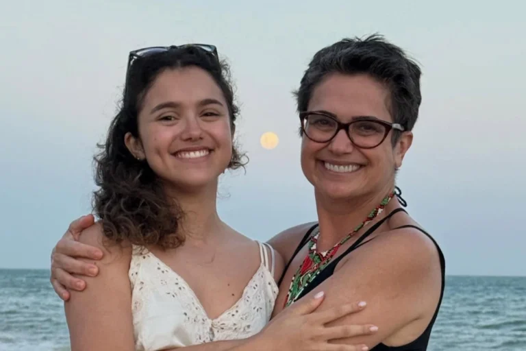 Sandra Annenberg e sua filha, Elisa Paglia, sorriem abraçadas em uma praia durante o entardecer. Sandra usa óculos de grau e um colar colorido, enquanto Elisa veste uma blusa branca de renda. Ao fundo, o mar e uma lua cheia no céu claro.