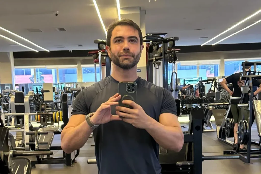Thiago Costa, filho do cantor Leandro, aparece em uma selfie de academia tirada no espelho. Ele está de pé, segurando um iPhone com as duas mãos. Ele veste uma camiseta preta ajustada e um relógio prateado no pulso esquerdo. Sua expressão é séria e focada. Ao fundo, vê-se um ambiente de academia moderno com diversas máquinas de musculação, anilhas, outras pessoas treinando e grandes janelas. A iluminação é brilhante e uniforme.