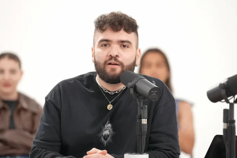 Tiago Santineli, humorista de barba e cabelo curto cacheado, falando ao microfone preto em um estúdio de podcast com fundo branco. Ele veste um moletom preto com estampa gráfica e usa colares de prata.