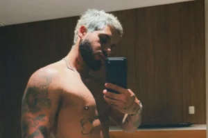 Zé Felipe sem camisa, com cabelo platinado e barba escura, tirando uma selfie no espelho. Ele exibe suas tatuagens no braço e no peito, incluindo um rosto sorridente e um desenho tribal. Ele está segurando um smartphone preto e usa um colar de corrente. O fundo é uma parede de madeira.