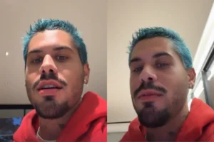 Cantor Zé Felipe com cabelo curto tingido de azul vibrante, usando um moletom vermelho com capuz. Ele possui barba e bigode aparados e olha diretamente para a câmera em um ambiente interno iluminado.