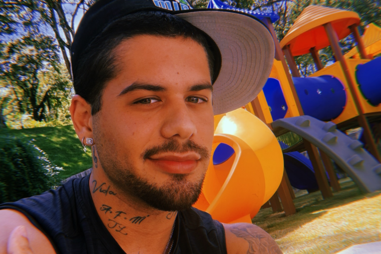 Zé Felipe em close, usando boné preto com aba cinza e regata preta. O cantor possui barba aparada, diversas tatuagens visíveis nos braços e pescoço, e posa em um ambiente externo com um parquinho infantil colorido ao fundo.