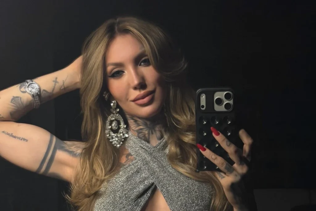 Texto Alternativo: A influenciadora Zoo posa para uma selfie em frente a um espelho, usando um top prateado brilhante com decote cruzado e um brinco de cristal grande e pendente. Ela tem cabelos loiros longos e ondulados, unhas vermelhas longas e exibe tatuagens detalhadas no pescoço e no braço esquerdo, além de um relógio prateado no pulso.