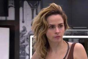 Close do rosto de Ana Paula Renault com expressão séria e atenta. Ela tem cabelos loiros e ondulados volumosos e usa uma regata marrom. O fundo é um ambiente de reality show com paredes de grafite e iluminação de neon.