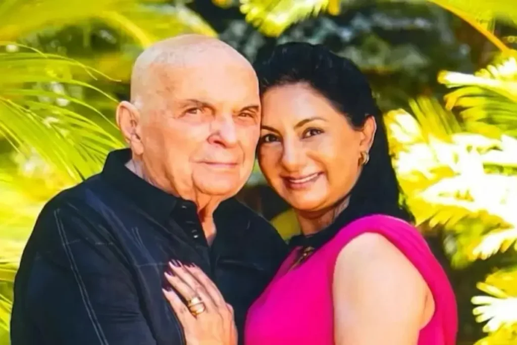 Retrato de Gerardo Henrique Machado, um senhor careca vestindo uma camisa preta, abraçado a Ana Gomes, uma mulher com cabelo escuro preso e blusa rosa choque. Os dois estão sorrindo e com os rostos colados, com um fundo verde de folhagens ensolaradas. A mão de Ana Gomes, com uma aliança dourada, está sobre o peito dele.
