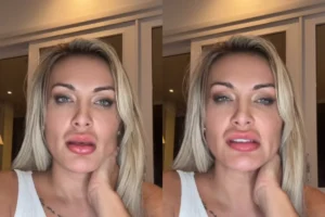 Mulher loira com olhos claros, vestindo uma regata branca, com a mão esquerda no pescoço e olhando seriamente para a câmera em um ambiente interno iluminado.