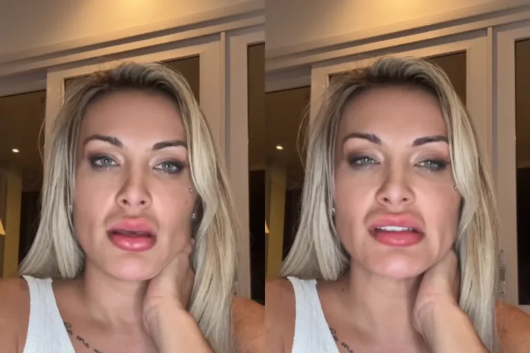 Mulher loira com olhos claros, vestindo uma regata branca, com a mão esquerda no pescoço e olhando seriamente para a câmera em um ambiente interno iluminado.