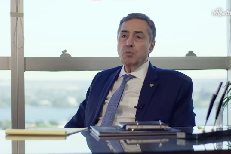 O ex-ministro e ex-presidente do STF, Luís Roberto Barroso, veste paletó azul-marinho e gravata clara. Ele aparece sentado em sua mesa de trabalho com vista para Brasília ao fundo.