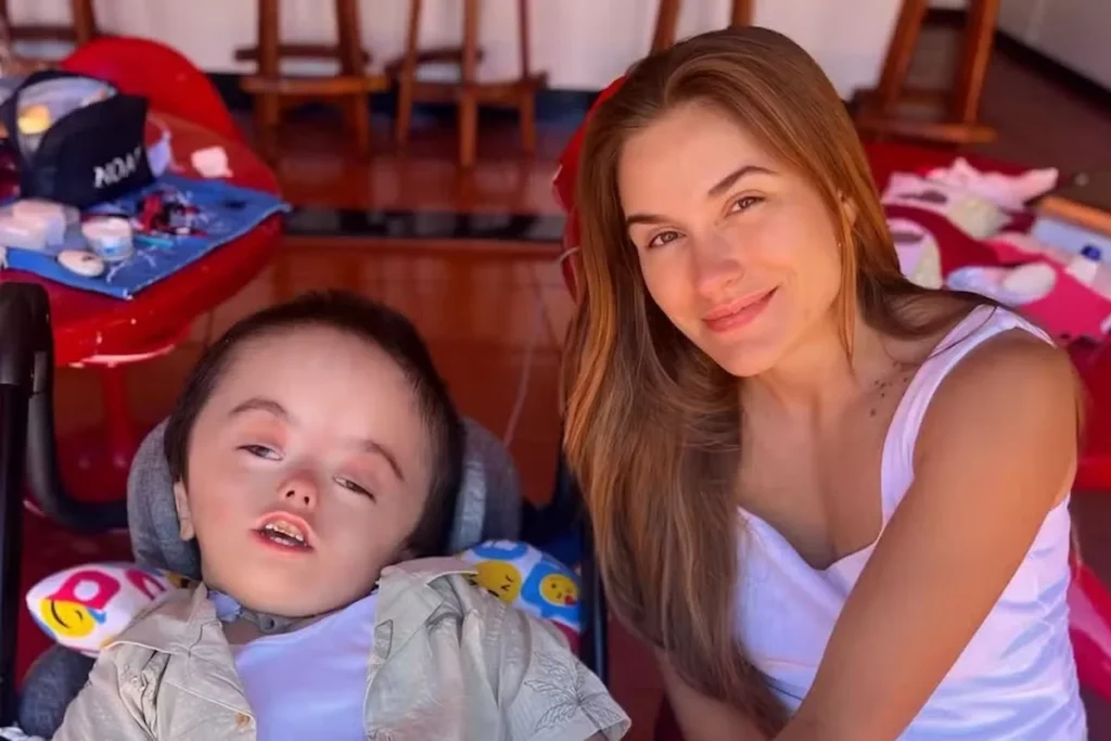 Bruna Massa, de cabelos castanhos claros e blusa branca, sorri ao lado de seu filho Noah, que está acomodado em uma cadeira adaptada com apoio cervical colorido.