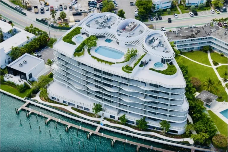 Prédio de luxo de arquitetura moderna e curvilínea em tons de branco e cinza, localizado à beira-mar. O terraço possui piscinas, áreas verdes com jardins suspensos e deck de madeira, com vista para o canal e barcos atracados.