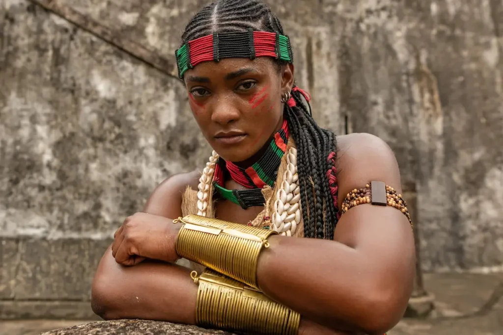 Erika Januza caracterizada com tranças nagô, faixa de miçangas coloridas na testa e pinturas vermelhas no rosto. Ela usa diversos acessórios como búzios, braceletes dourados largos e colares étnicos, posando com os braços cruzados diante de um muro de pedra.