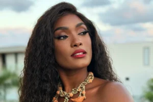 Erika Januza posa de perfil com cabelos longos e ondulados, usando maquiagem marcante com batom vermelho e um colar de contas rústicas em tons terrosos. O fundo mostra um céu claro ao entardecer e parte de uma estrutura urbana.