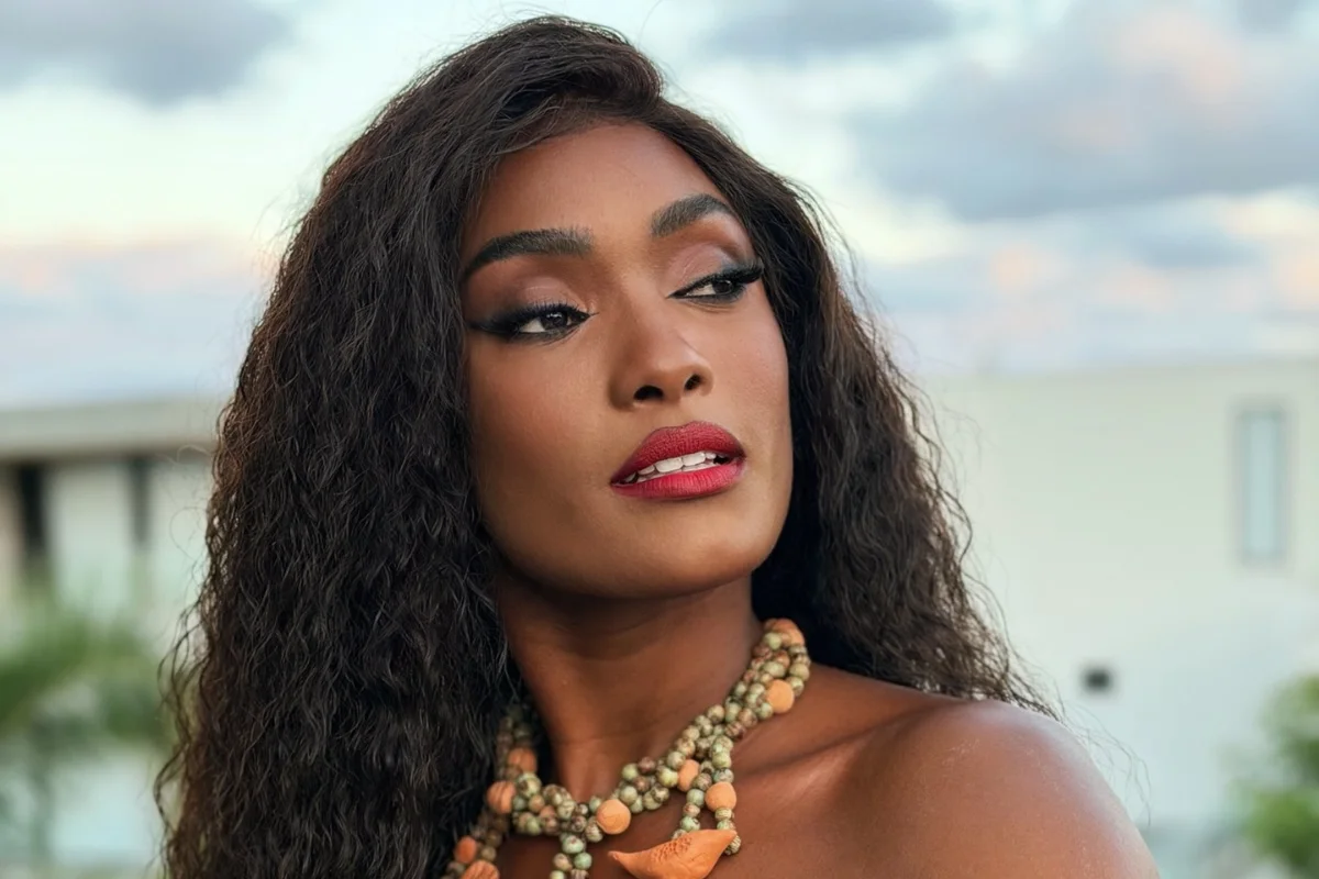 Erika Januza posa de perfil com cabelos longos e ondulados, usando maquiagem marcante com batom vermelho e um colar de contas rústicas em tons terrosos. O fundo mostra um céu claro ao entardecer e parte de uma estrutura urbana.