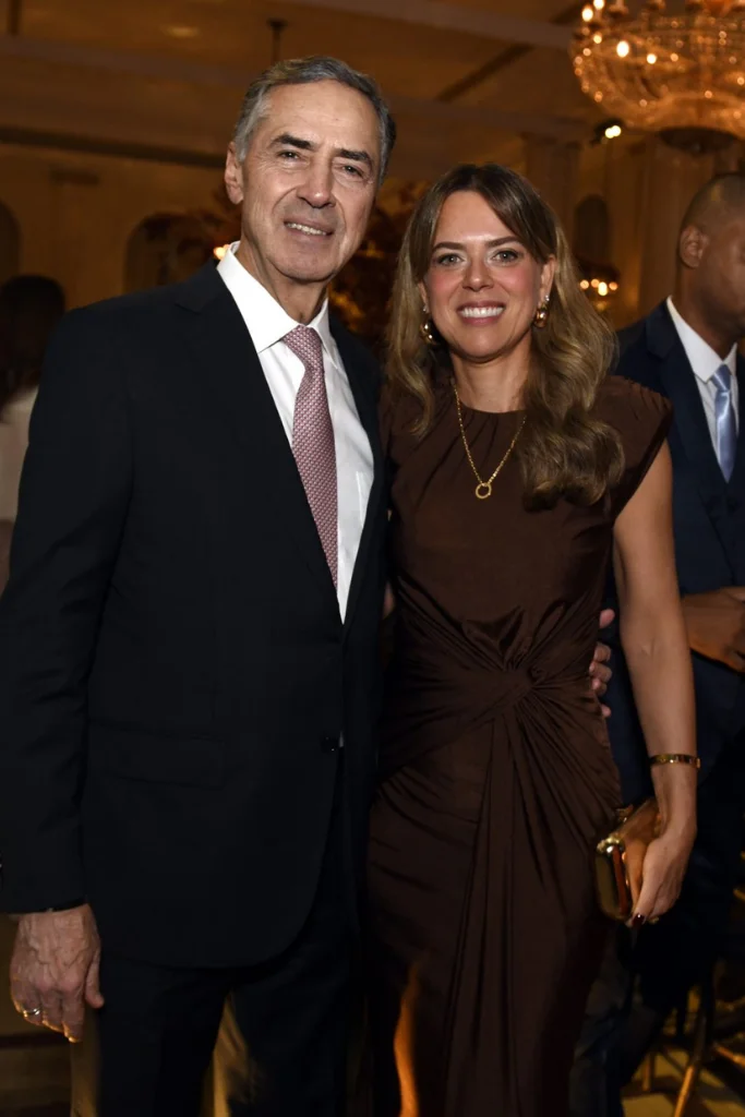 O ex-ministro Luís Roberto Barroso, em terno escuro e gravata rosa, sorri ao lado de sua namorada, Rita Nolasco, que usa um vestido marrom drapeado e segura uma bolsa clutch dourada em um evento social elegante.
