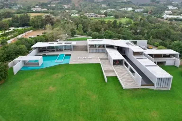 Vista aérea de uma mansão luxuosa de arquitetura brutalista em concreto aparente, com linhas retas e amplas. A propriedade conta com uma piscina de borda infinita em formato geométrico, um extenso gramado verde impecável e é cercada por colinas e vegetação densa.