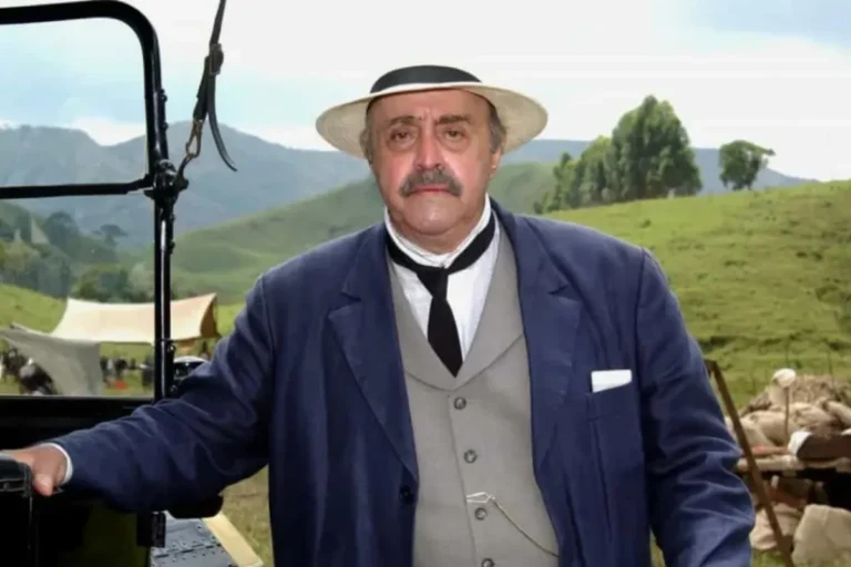 O ator Mauro Mendonça caracterizado com figurino de época, vestindo terno azul, colete cinza, gravata preta e chapéu de palha, posando em uma paisagem rural com montanhas ao fundo.