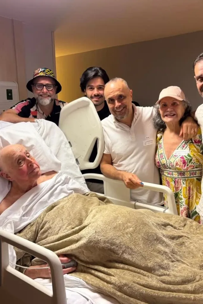 Mauro Mendonça deitado em um leito de hospital, sorrindo levemente, cercado por cinco familiares e amigos que estão em pé ao redor da cama, todos sorridentes.