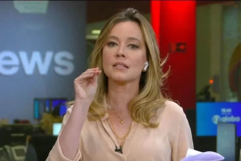 Jornalista Natuza Nery em estúdio de televisão, gesticulando com a mão direita enquanto fala. Ela possui cabelos loiros ondulados, usa uma blusa bege de seda, colares dourados finos e fones de ouvido sem fio brancos. Ao fundo, vê-se parte do cenário da GloboNews com monitores e iluminação técnica.