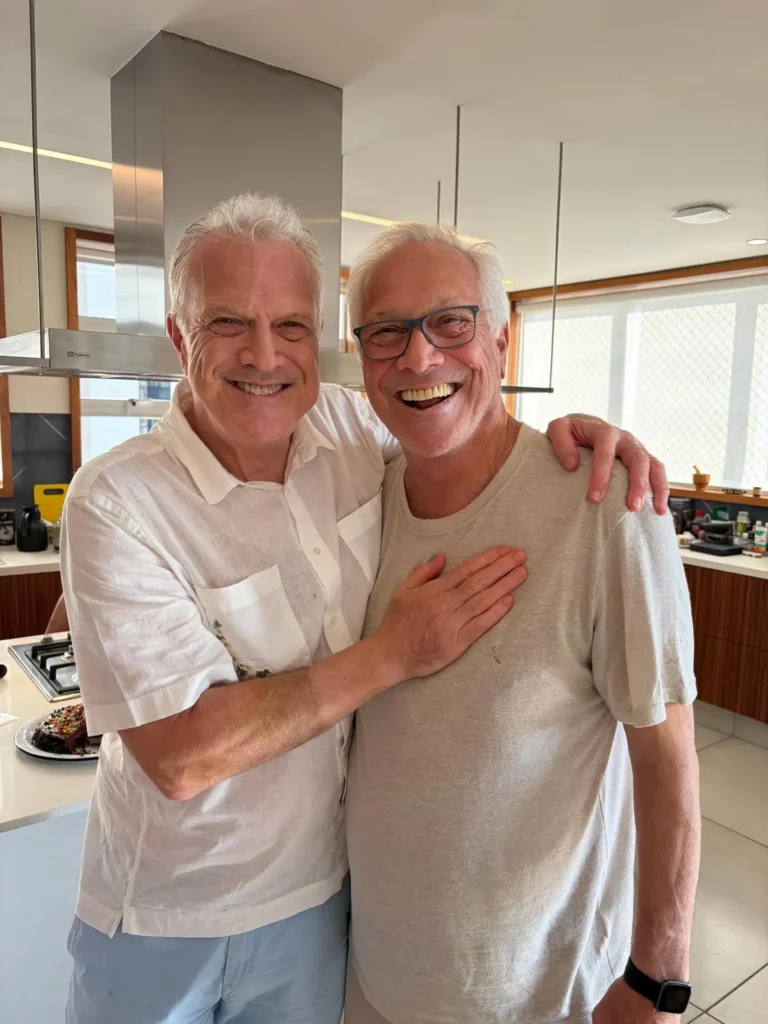 Pedro Bial e seu irmão Alberto Bial sorridentes, abraçados em uma cozinha de decoração moderna. Pedro Bial veste uma camisa social branca de mangas curtas e Alberto usa uma camiseta bege e óculos de armação escura.