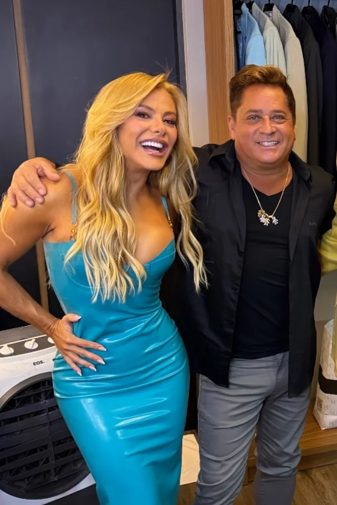 Poliana Rocha, loira com vestido justo azul brilhante, sorri ao lado do cantor Leonardo, que veste camisa preta e calça cinza; ambos aparecem abraçados em um closet com roupas ao fundo.