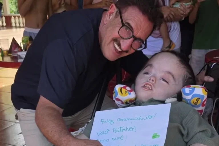 O apresentador Ratinho sorri inclinado ao lado de seu neto, Noah, que está em sua cadeira de rodas. Noah segura um cartaz feito à mão com os dizeres "Feliz Aniversário Vovô Ratinho! Amo você!". Ratinho usa óculos de armação preta e camisa escura.