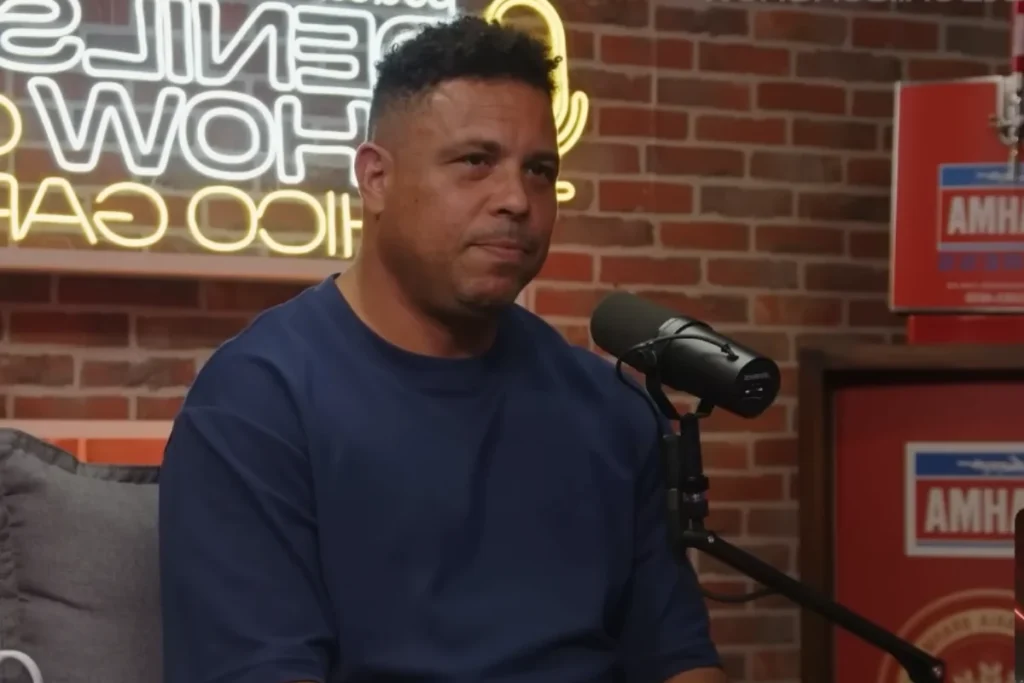 Ronaldo Fenômeno sentado de perfil em um estúdio de podcast, usando uma camiseta azul marinho. Ao fundo, uma parede de tijolos expostos com um letreiro luminoso em neon amarelo e um microfone profissional à sua frente.