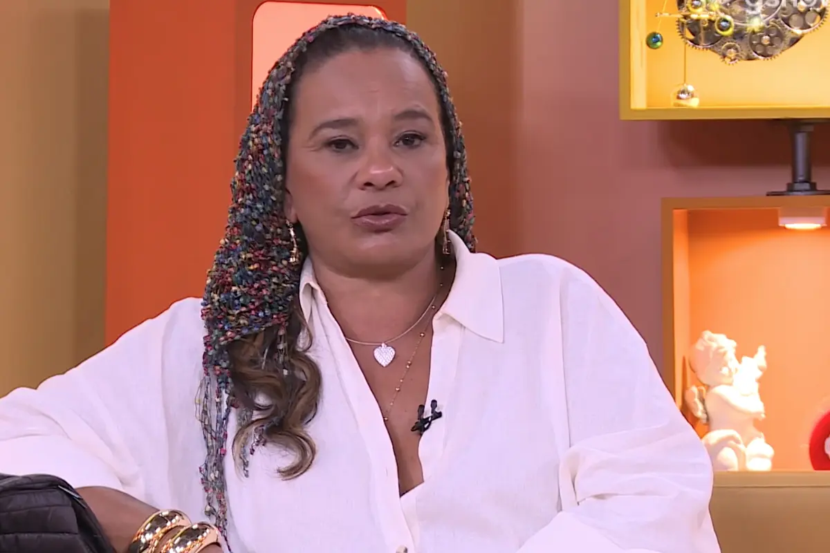 Atriz Solange Couto sentada em um sofá, vestindo uma camisa branca de botões, colares dourados com pingente de coração e pulseiras largas. Ela utiliza um lenço colorido tramado sobre a cabeça e o ombro. O fundo apresenta elementos decorativos em tons de laranja e amarelo.