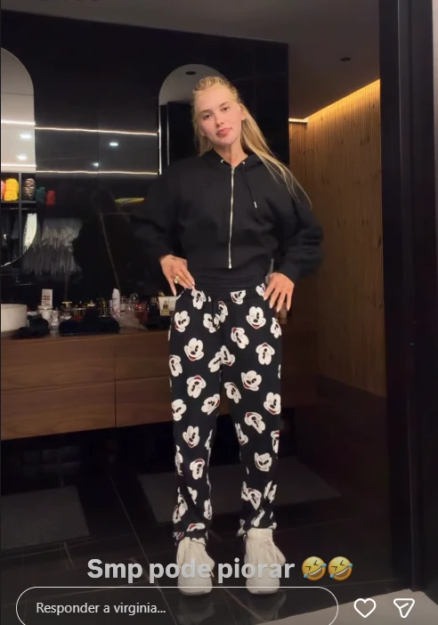 Virginia Fonseca posa em pé vestindo um casaco de moletom preto com zíper e calça preta estampada com o rosto do personagem Mickey Mouse. Ela está em um ambiente interno, possivelmente um closet, com iluminação suave.