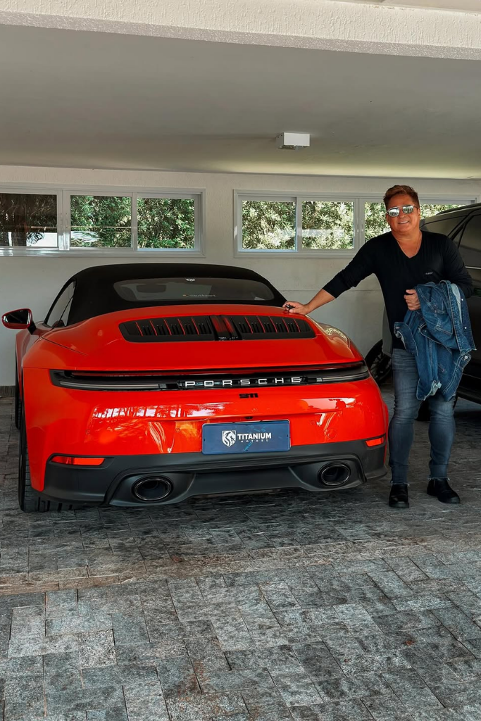 Cantor Leonardo sorridente em uma garagem, vestindo roupa preta e óculos escuros, apoiando a mão na traseira de um Porsche 911 conversível na cor vermelha. O carro de luxo possui capota preta e placa da Titanium Motors.