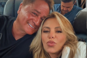 Close do rosto de Leonardo e Poliana Rocha; ele sorri encostando a cabeça na dela, enquanto Poliana faz um biquinho para a selfie. Ambos estão no que parece ser o interior de uma aeronave ou ônibus de viagem.