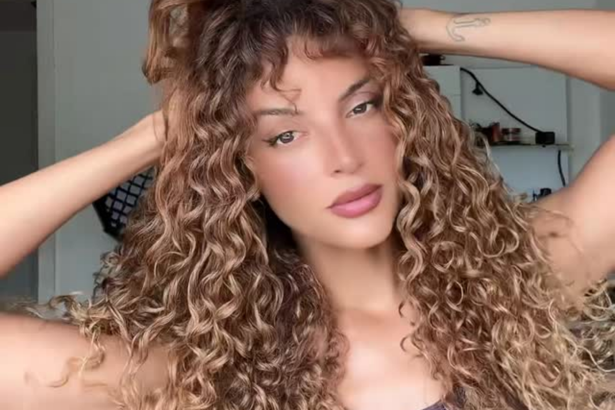 Close de Gabriela Loran olhando para a câmera e com as mãos no cabelo. Ela exibe cabelos cacheados volumosos e loiros, batom rosado e delineado nos olhos. Uma pequena tatuagem de âncora é visível no pulso direito. O fundo é de um ambiente interno com prateleiras desfocadas.