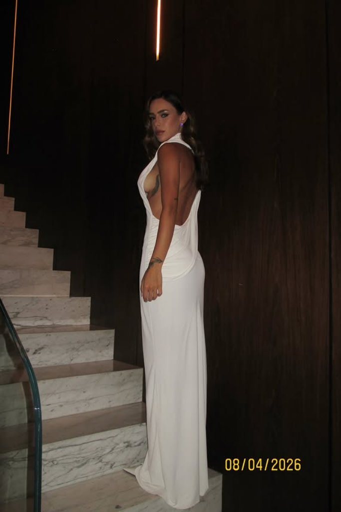 Bruna Biancardi posa de perfil em uma escadaria de mármore, olhando para trás. Ela usa um vestido longo branco justo, sem mangas e com as costas totalmente abertas, revelando uma tatuagem de pena na costela. O ambiente tem paredes de madeira escura e iluminação focada. Uma data amarela