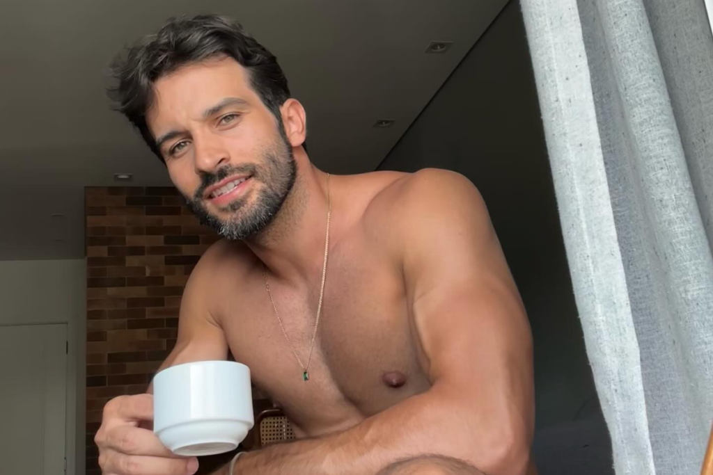 Close-up de Bruno Bevan, sem camisa, sorrindo e segurando uma xícara branca de café. Ele tem uma barba bem aparada e usa um colar dourado. O fundo mostra um ambiente interno, com uma parede de tijolos e uma cortina clara.