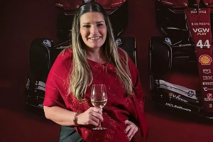 Bianca Naufel Saliba sorrindo, vestindo uma echarpe vermelha sobre roupa escura, segurando uma taça de vinho branco. Ao fundo, em um ambiente de iluminação cênica, destaca-se um carro de Fórmula 1 da equipe Ferrari com o número 44.