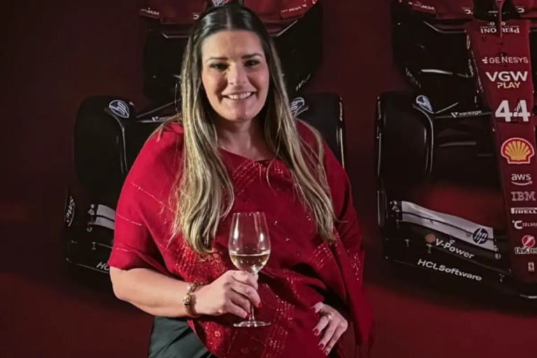 Bianca Naufel Saliba sorrindo, vestindo uma echarpe vermelha sobre roupa escura, segurando uma taça de vinho branco. Ao fundo, em um ambiente de iluminação cênica, destaca-se um carro de Fórmula 1 da equipe Ferrari com o número 44.