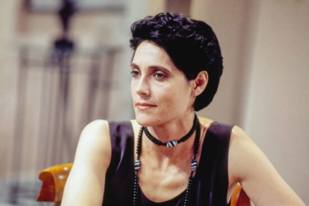 Christiane Torloni como a personagem Diná na novela A Viagem, com cabelos curtos e escuros, vestindo blusa preta e colares de contas, mantendo uma expressão pensativa.