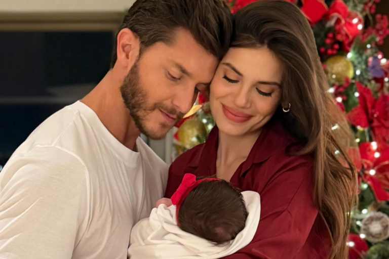Klebber Toledo e Camila Queiroz aparecem abraçados e sorridentes enquanto seguram a filha recém-nascida, Clara, que usa um laço vermelho na cabeça. Ao fundo, vê-se uma árvore de Natal decorada com luzes e ornamentos vermelhos.