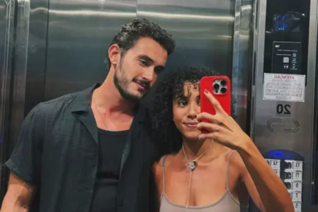 Filipe Bragança e Ramille posam para selfie em frente ao espelho de um elevador. Ramille segura um celular com capa vermelha; ela tem cabelos cacheados e usa uma blusa de alças finas. Filipe está ao lado dela, vestindo uma camisa preta aberta sobre uma regata escura.