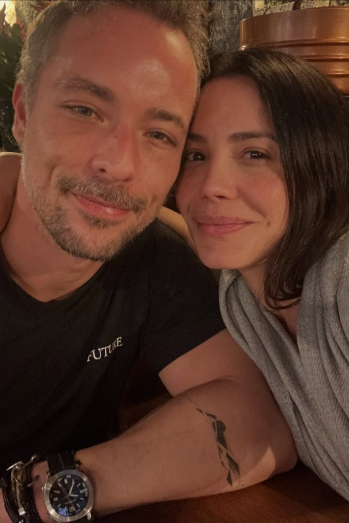 Bernardo Virtuoso e Juliana Knust sorrindo abraçados em um ambiente aconchegante. Bernardo veste camiseta preta com a palavra