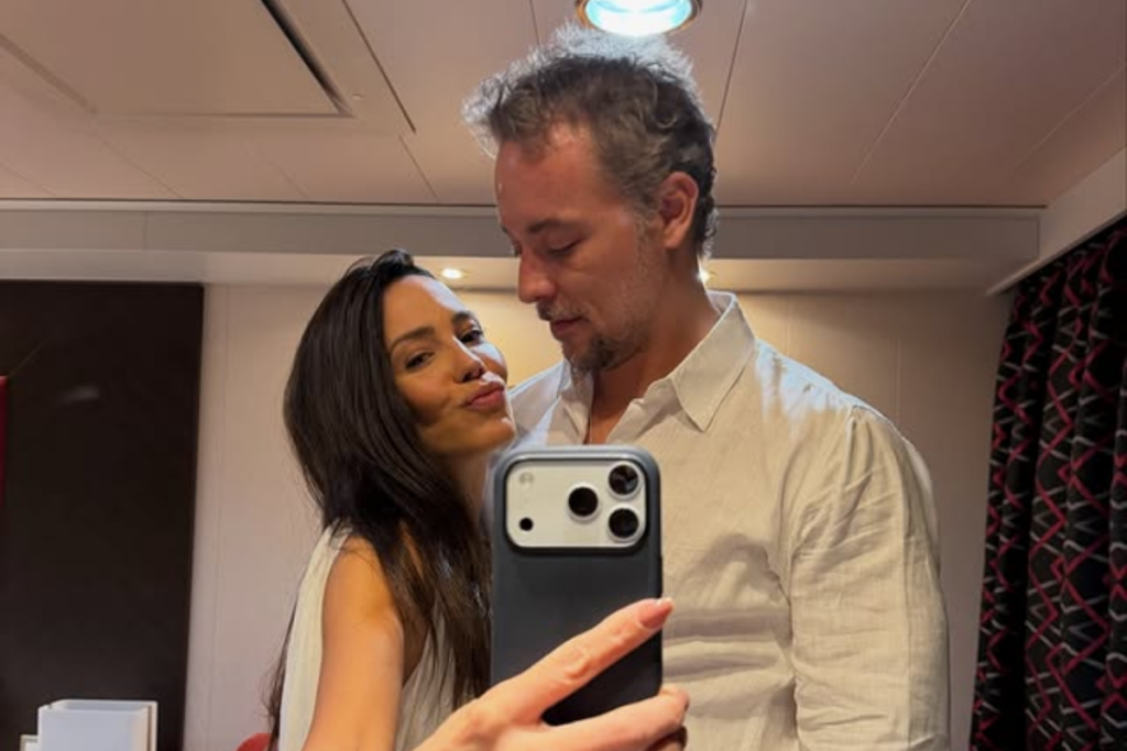 Juliana Knust fazendo biquinho e Bernardo Virtuoso olhando para ela em uma selfie no espelho. Juliana está à esquerda com cabelos longos e Bernardo veste uma camisa social branca à direita.