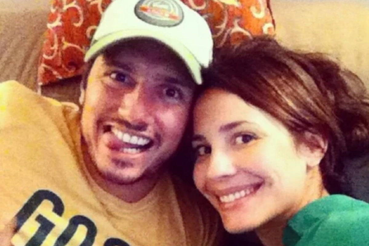 Close selfie de Juliana Knust e Gustavo Machado. Gustavo, à esquerda de boné cinza e amarelo, sorri abertamente e mostra a língua. Juliana, à direita, sorri amplamente e encosta a cabeça na dele. Eles estão em um ambiente interno com um travesseiro com estampa arabesca laranja ao fundo.